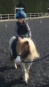 James riding a pony in an outdoor arena. The horse is being lead by someone off camera.
