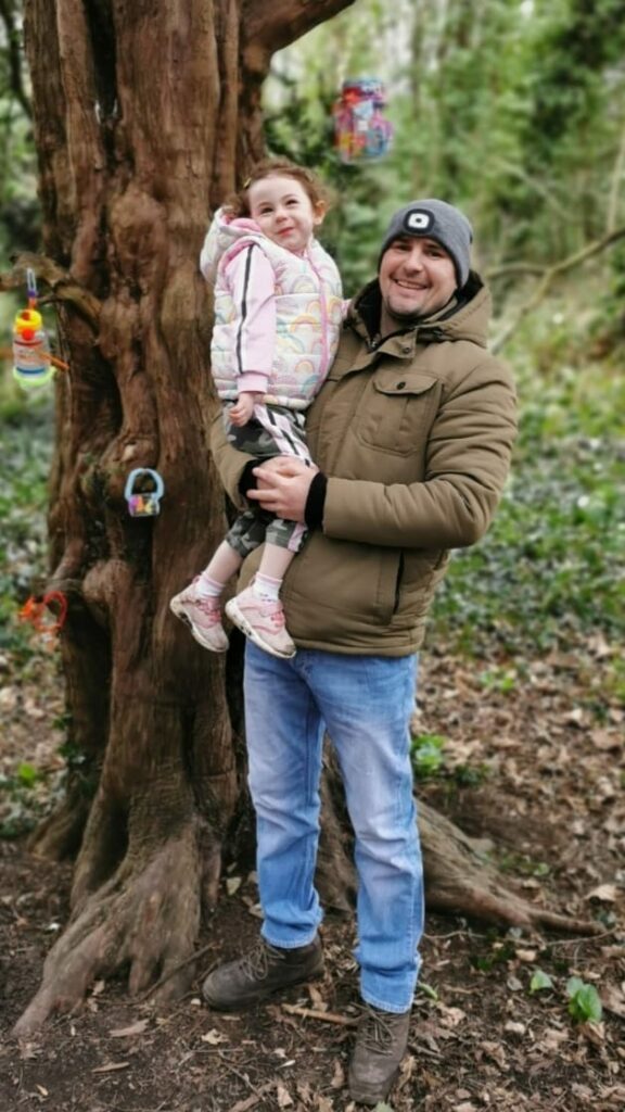Bonnie's dad is holding her outside by a tree, they are both smiling at the camera