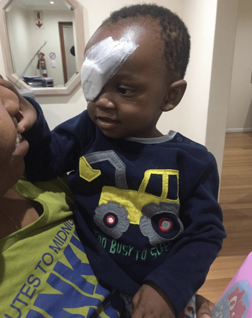 Joseph has a bandage over his eye. He is looking at his mum as she cuddles him smiling