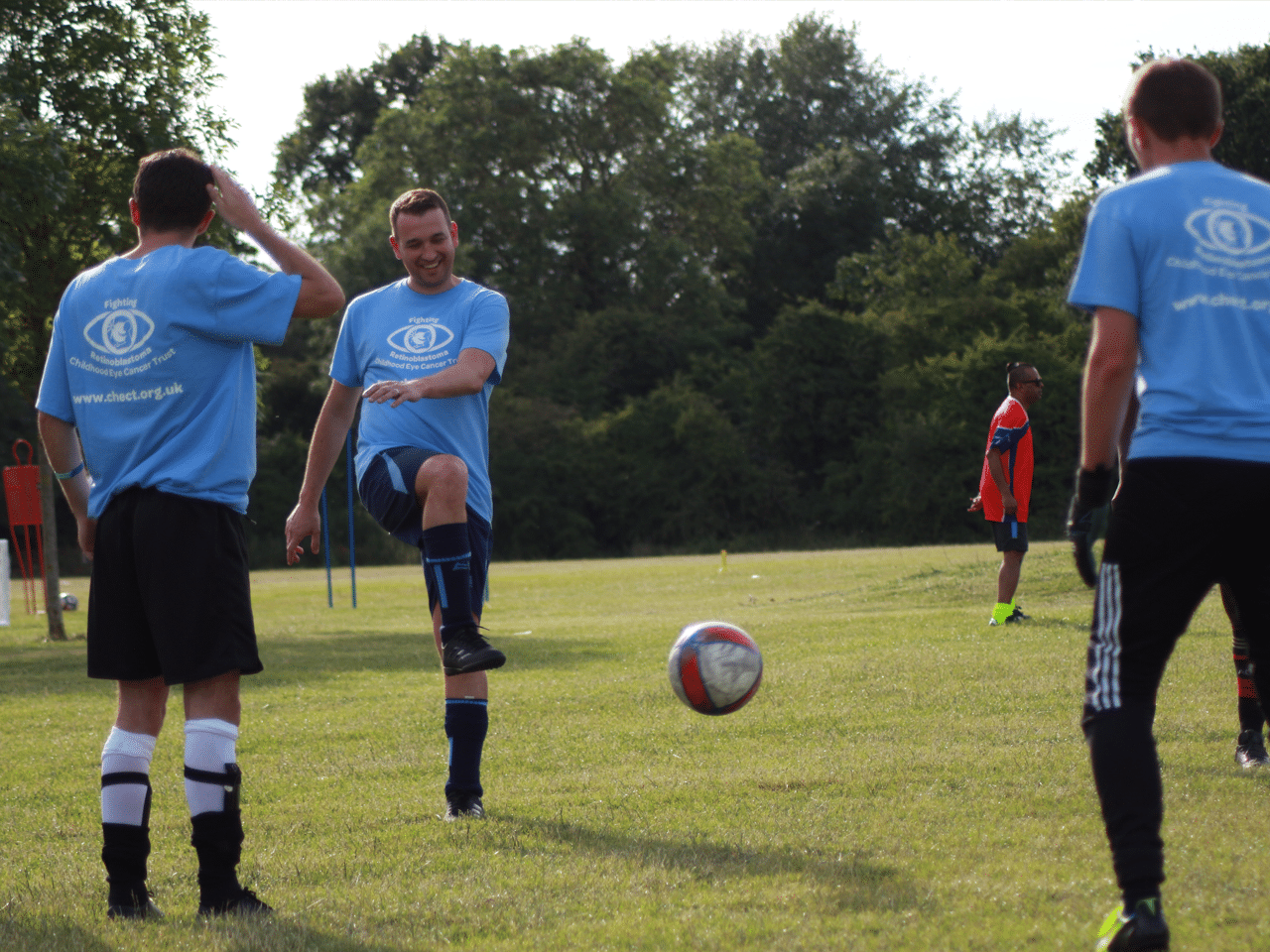 Footy football being kicked by childhood eye cancer trust member towards two of his friends, all in chect branded t-shrits