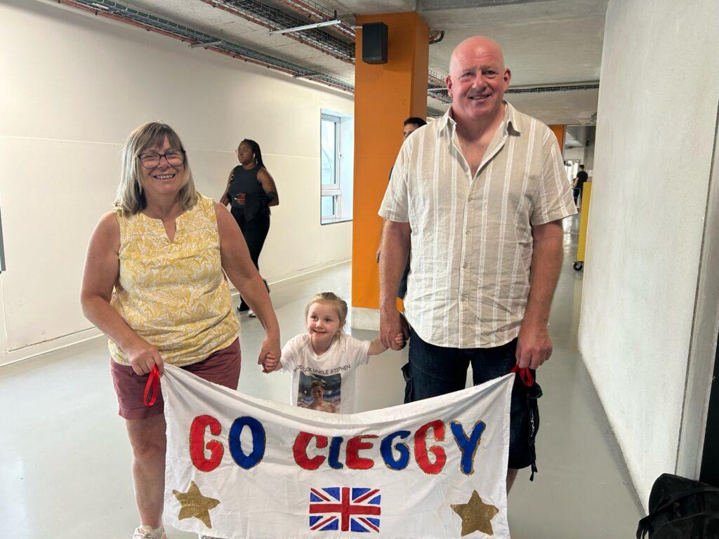 Adaline is stood with her grandparents holding a handmade 'Go Cleggy' sign