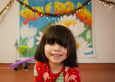 Little girl in the centre of the picture with a short brown bob is smiling straight at the camera. She's wearing a Christmassy dress and in the background are Christmas decorations - very festive!