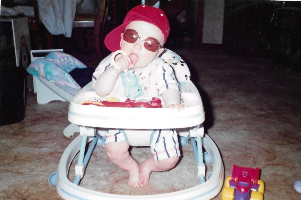 Adam as a baby wearing sunglasses and a backwards cap in a baby chair