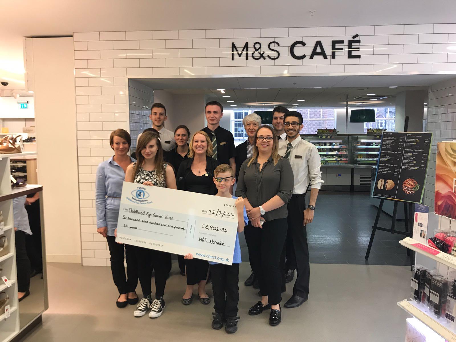 RS2156_M&S Norwich presentation M&S colleagues and childhood eye cancer trust family standing in front of the M&S cafe in Norwich holding a big cheque!
