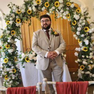 Danilo is smiling in a wedding suit, with a floral arch behind him